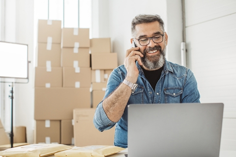 man speaking on the phone and looking at his computer with boxes behind him